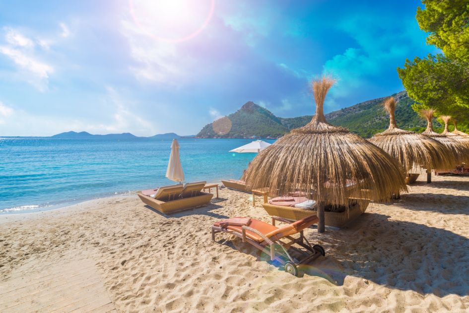 View over the crystal clear waters of Formentor Beach in Palma de Mallorca, with its sandy beach and sun lounger in the foreground. A great spot for luxury holidays in the Balearics.