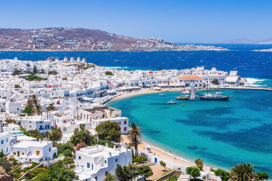 View over the whitewashed buildings of Mykonos, one of the best luxury holiday destination. The crystal clear waters of the Aegean Sea are set in the foreground and the hillside landscape in the background. 