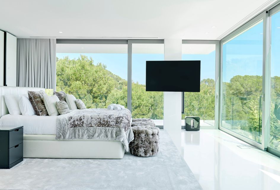 Panoramic view of Villa La Costa's master bedroom, featuring a flat screen TV and floor to ceiling windows, offering a stunning natural background. A top choice for a secluded villa holiday in Ibiza.