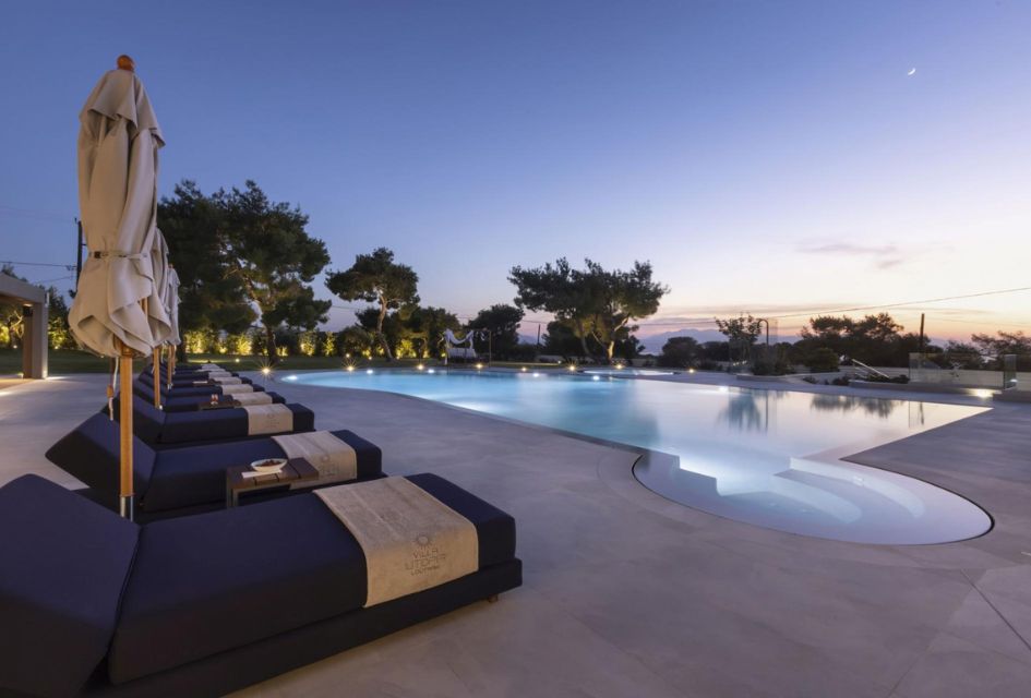 The large luxury swimming pool of Villa Utopia at dusk, with the crescent moon in the sky and deep blue loungers giving an elegant touch to the outside area. Lights shine around the pool of this Peloponnese villa.